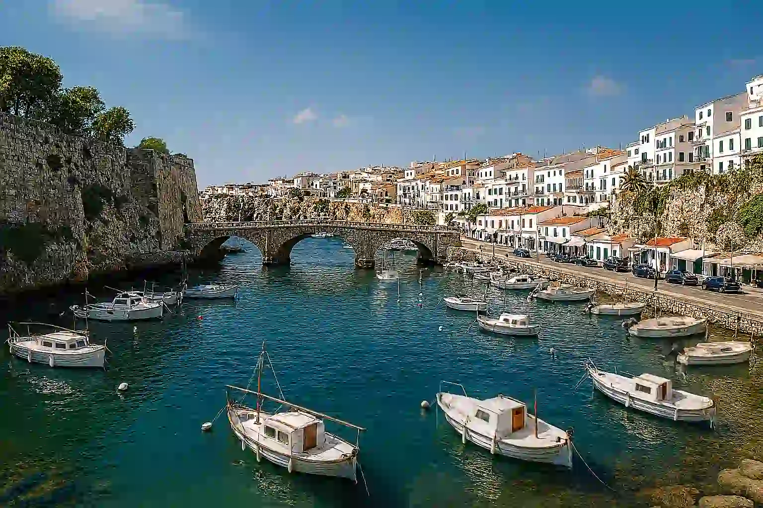 Cliff-framed cove in Menorca with turquoise water, stone paths and a whitewashed village